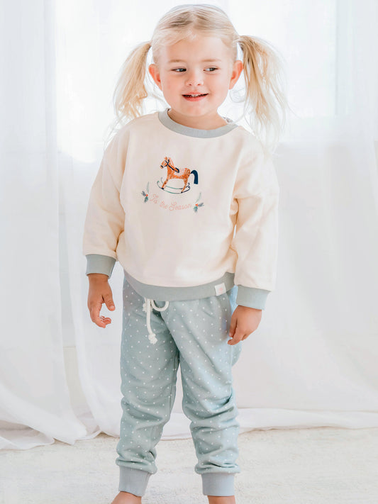 This image of a girl features the product Sweatshirt Set - O Christmas Tree. The light blue top features an illustration of an old rocking horse and the words ‘Tis the Seaon’ in light pink. The bottoms are a pattern of white polka dots on a dusty light blue.