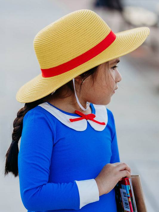 This image of a girl features the product Straw Hat - Madeline. This bright yellow wide brimmed hat has a red stripe around it and is the perfect finishing touch for a Madeline costume when paired with our Madeline Swing Dress. 